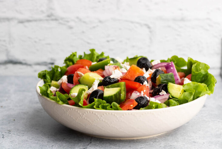 A white bowl filled with chopped tomatoes, peppers, red onions, cucumbers, black olives, and feta cheese.