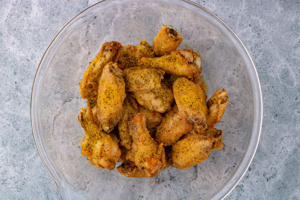 A glass bowl filled with par baked chicken wings covered in Lemon Pepper honey butter.