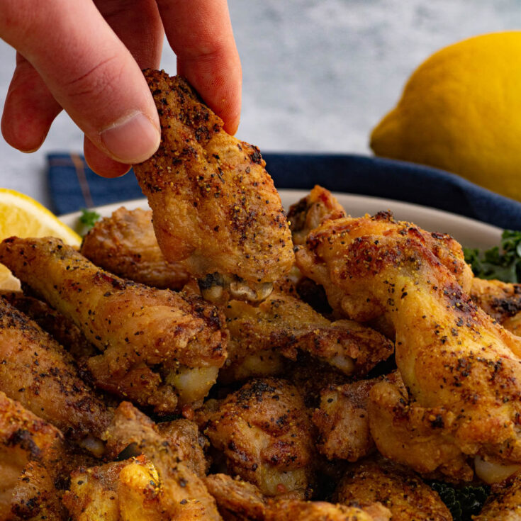 Fingers grabbing a single baked lemon pepper wing out of a filled bowl.