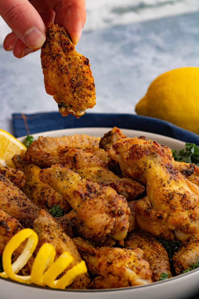 Fingers holding a single baked lemon pepper wing above a filled bowl.