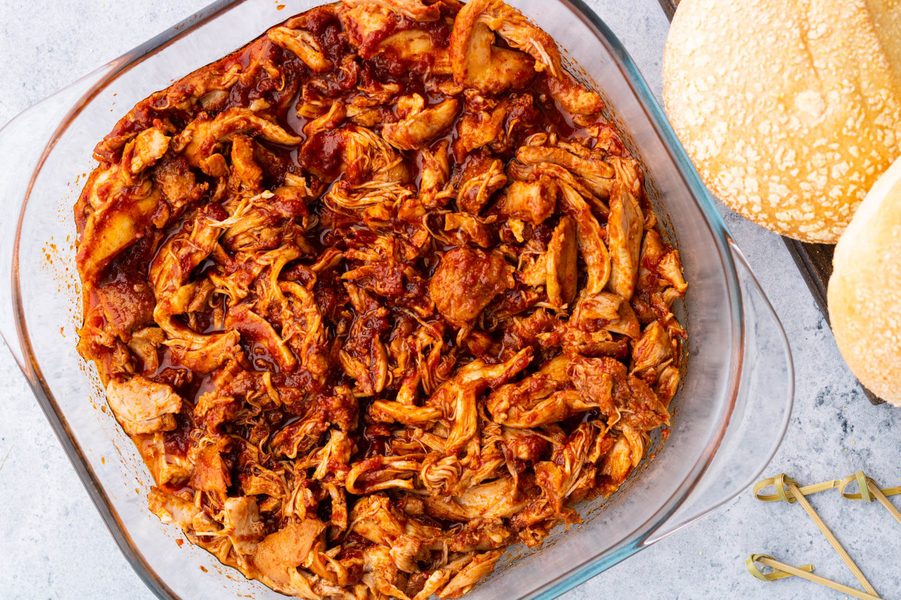 A glass baking dish containing BBQ Pulled Chicken beside buns.