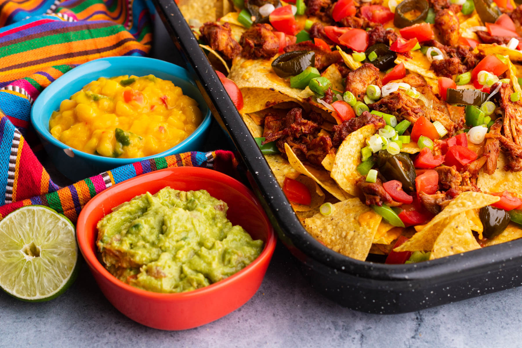 A colourful tray of BBQ Chicken nachos with two small side dishes of mango salsa and guacamole.