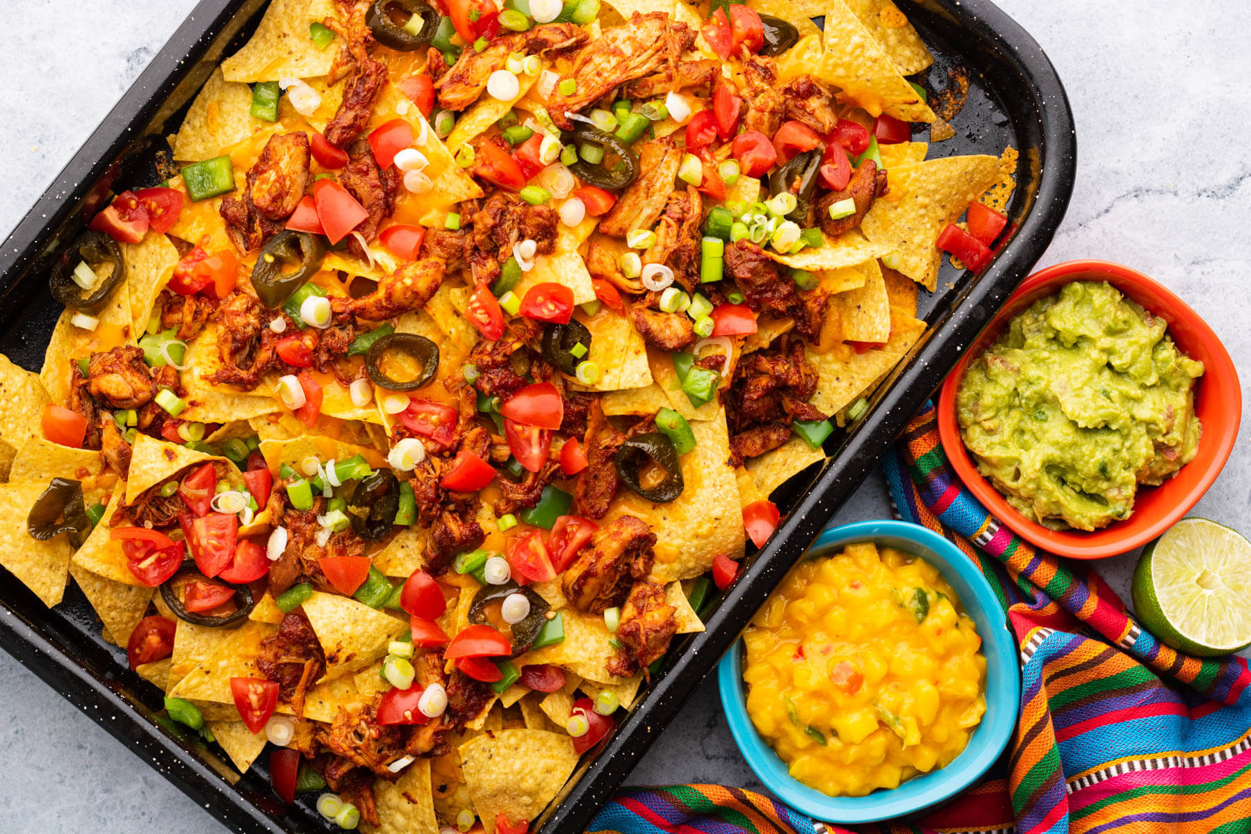 A colourful tray of BBQ Chicken nachos with two small side dishes of mango salsa and guacamole.