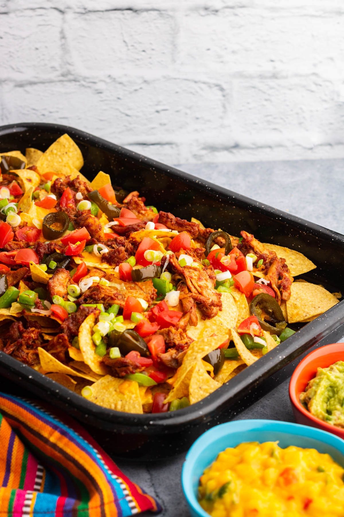A colourful tray of BBQ Chicken nachos with two small side dishes of mango salsa and guacamole.