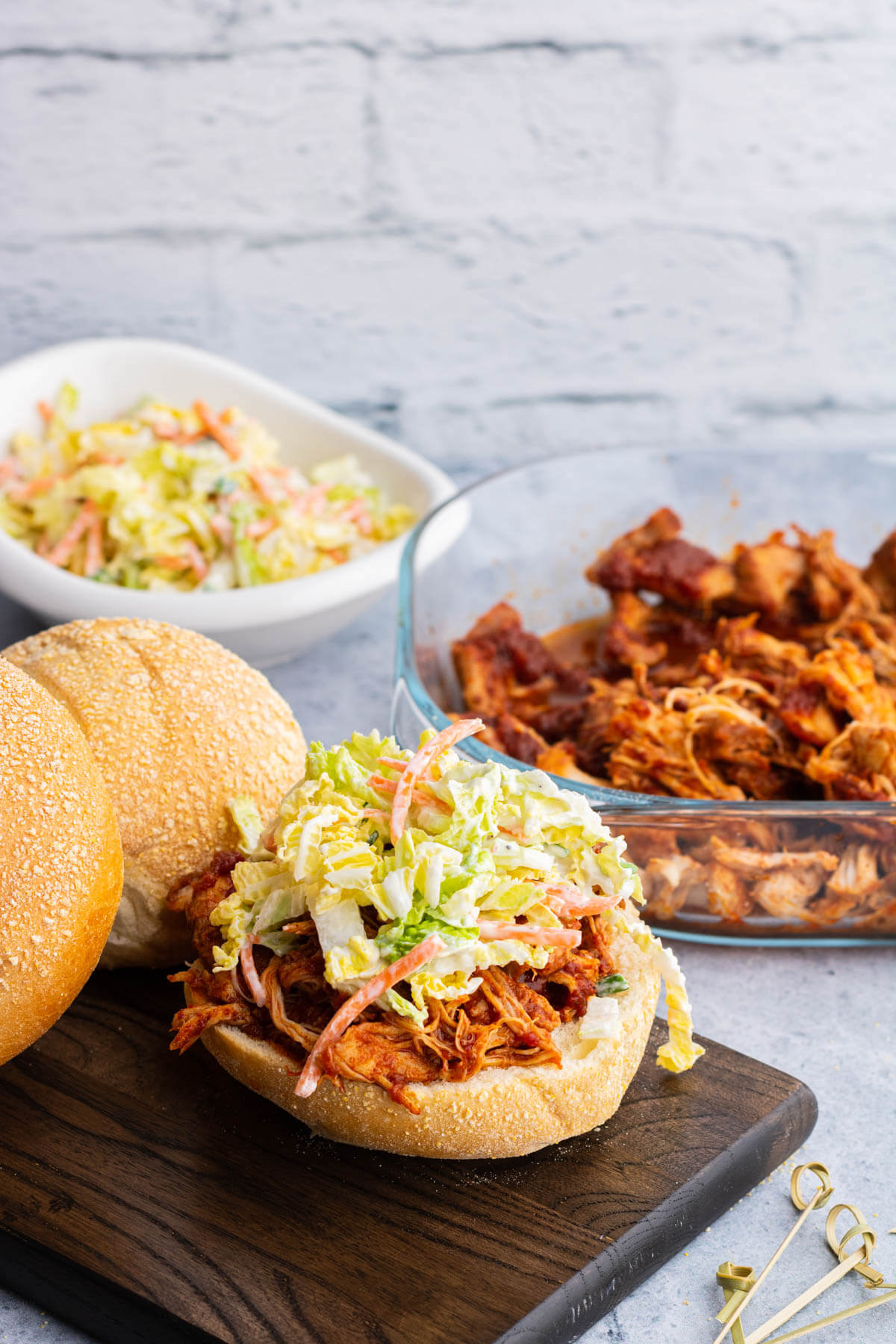 A soft white bun containing BBQ Pulled Chicken and coleslaw surrounded by a glass dish of BBQ Pulled Chicken, coleslaw, and buns.