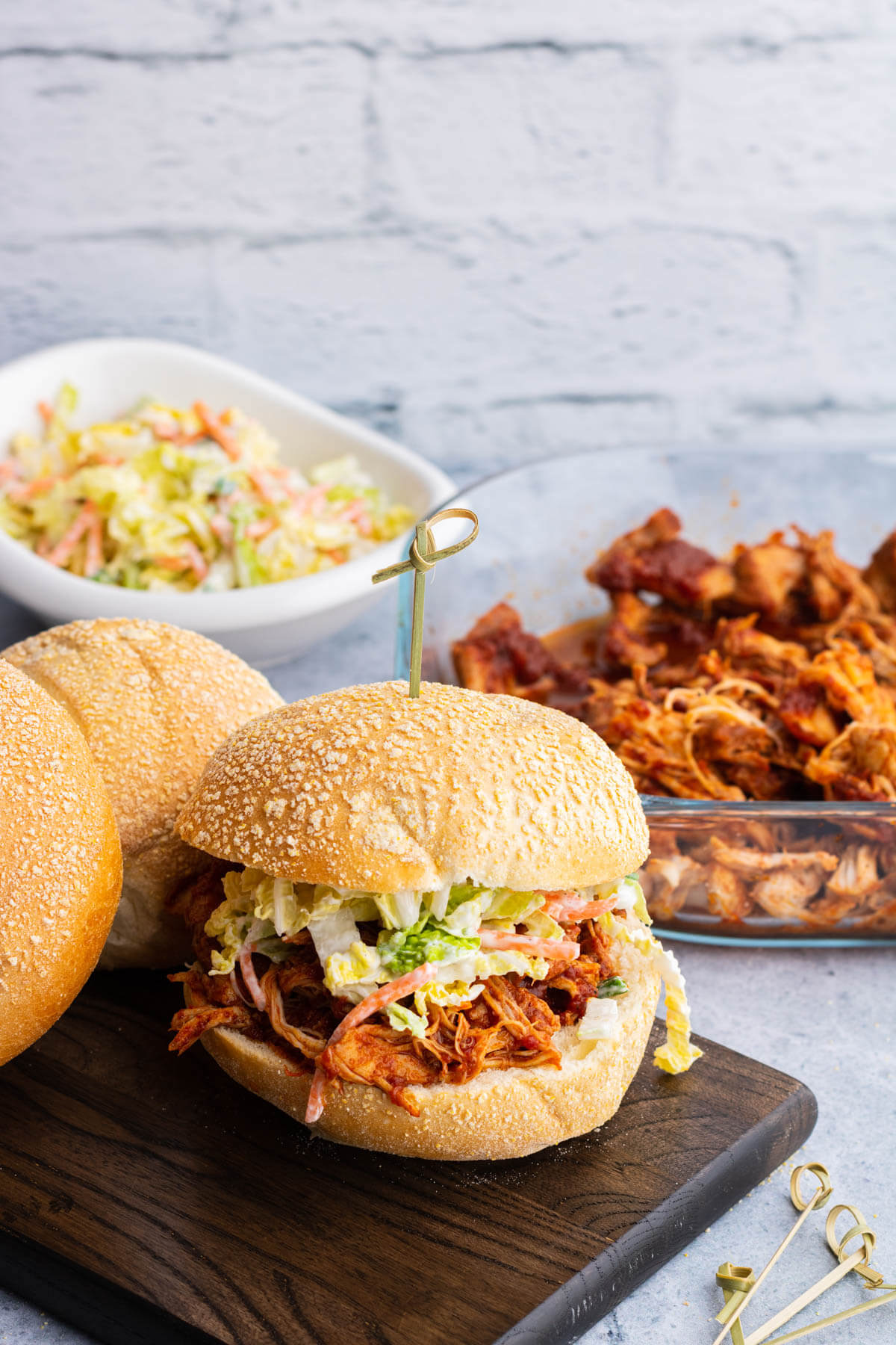 A soft white bun containing BBQ Pulled Chicken and coleslaw surrounded by a glass dish of BBQ Pulled Chicken, coleslaw, and buns.