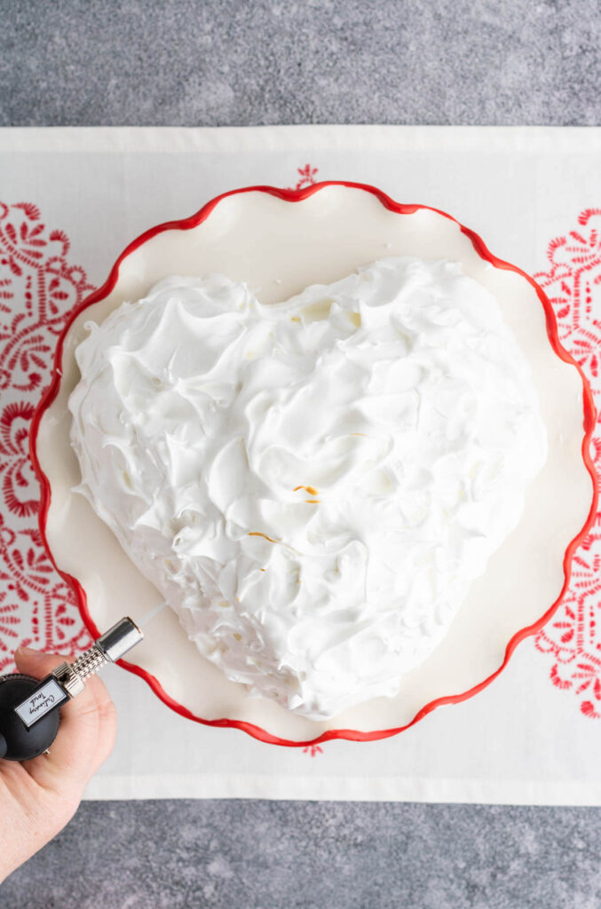 A heart shaped Baked Alaska covered in white meringue getting torched with a culinary torch.