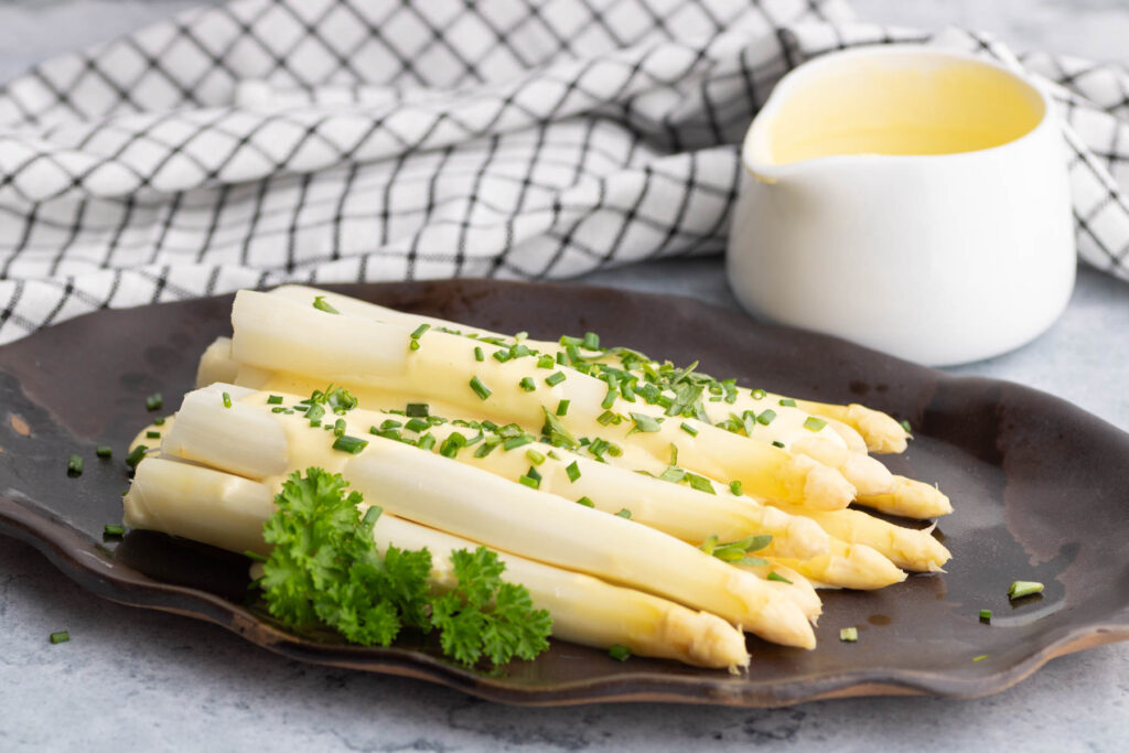Steamed white asparagus covered in pale yellow hollandaise and green herbs.