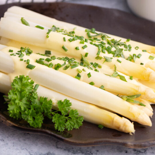 Steamed white asparagus covered in pale yellow hollandaise and green herbs.