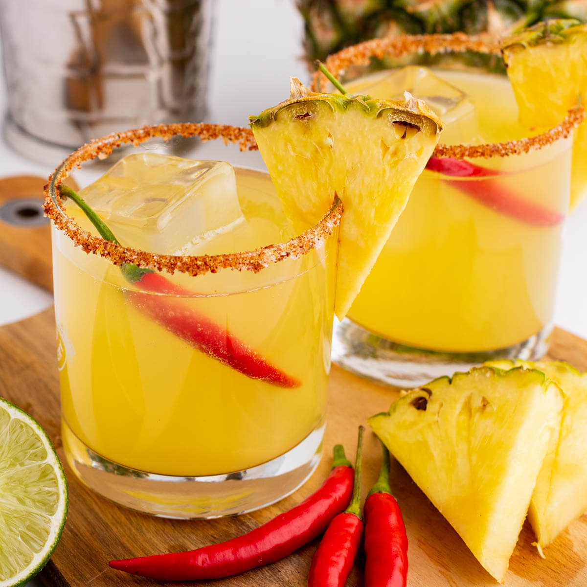 Two Spicy Pineapple Margaritas in red rimmed rocks glasses garnished with a wedge of pineapple and red chili pepper.