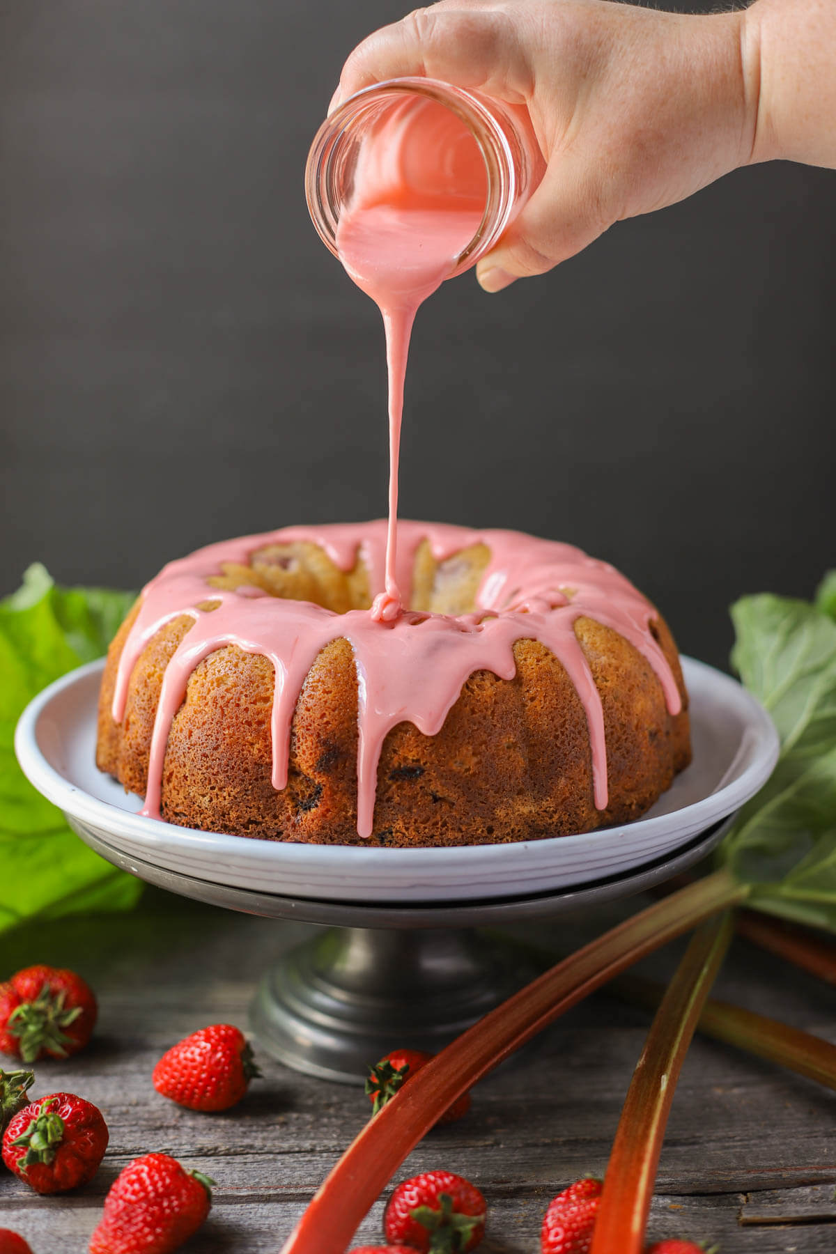 A hand pours pink glaze out of a jar over a golden baked bundt cake.
