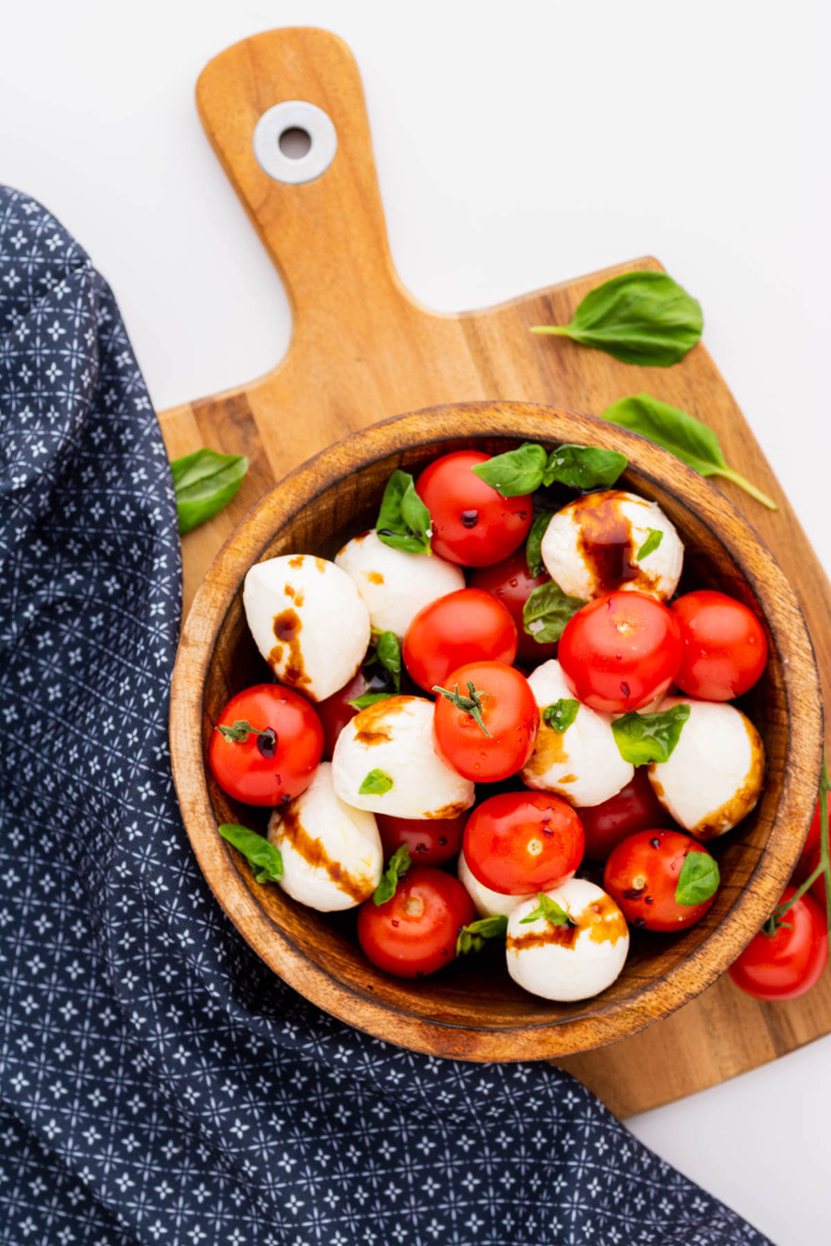 A wooden bowl filled with bright red cherry tomatoes, white mozzarella balls and green basil drizzled with olive oil and balsamic reduction.