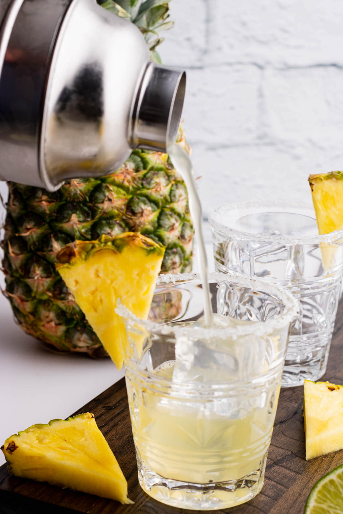 A cocktail being poured into a waiting rimmed rocks glass filled with ice and garnished with a slice of pineapple.