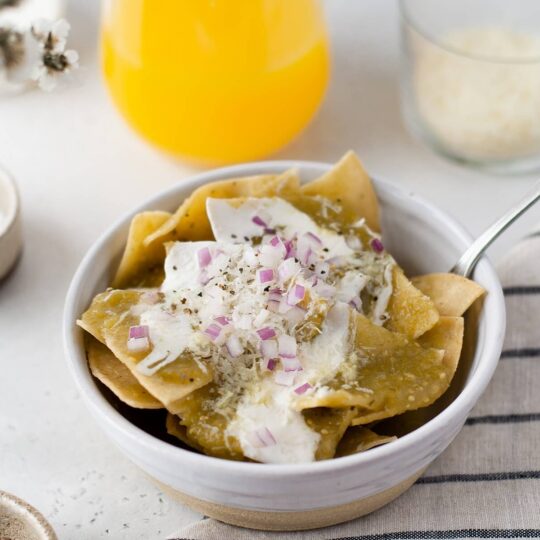 A breakfast table setting featuring a bowl of Chilaquiles Verdes topped with sour cream, diced purple onions, cilantro, and cotija cheese.