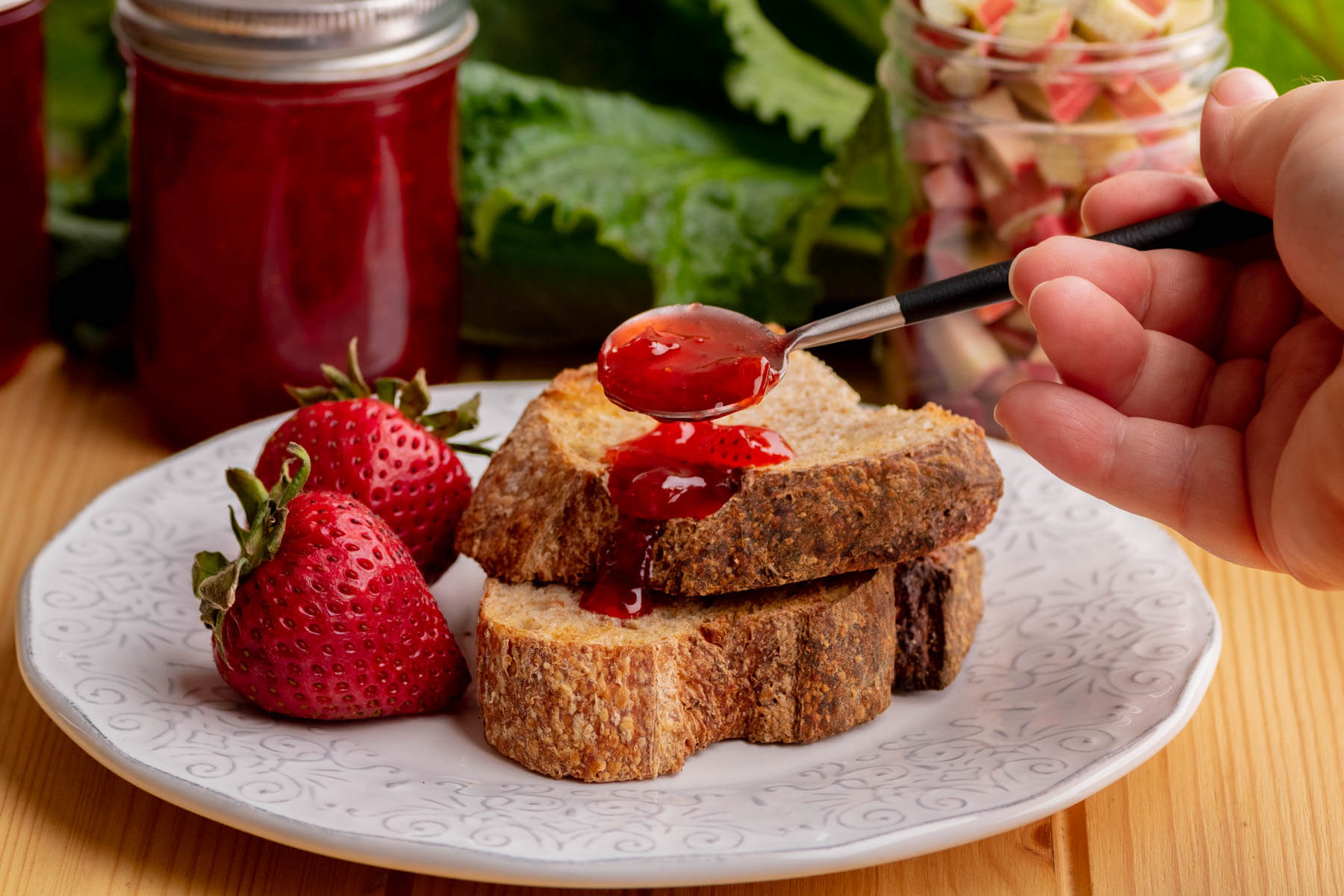 Two sliced of toast slathered in Strawberry Rhubarb Jam with a jar of jam and fresh rhubarb in the background.