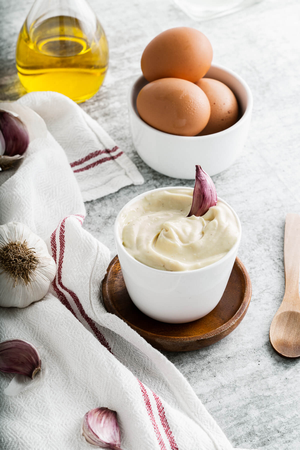 A small white dish filled with creamy Homemade Aioli garnished with a purple clove of garlic. Surrounded by aioli ingredients.