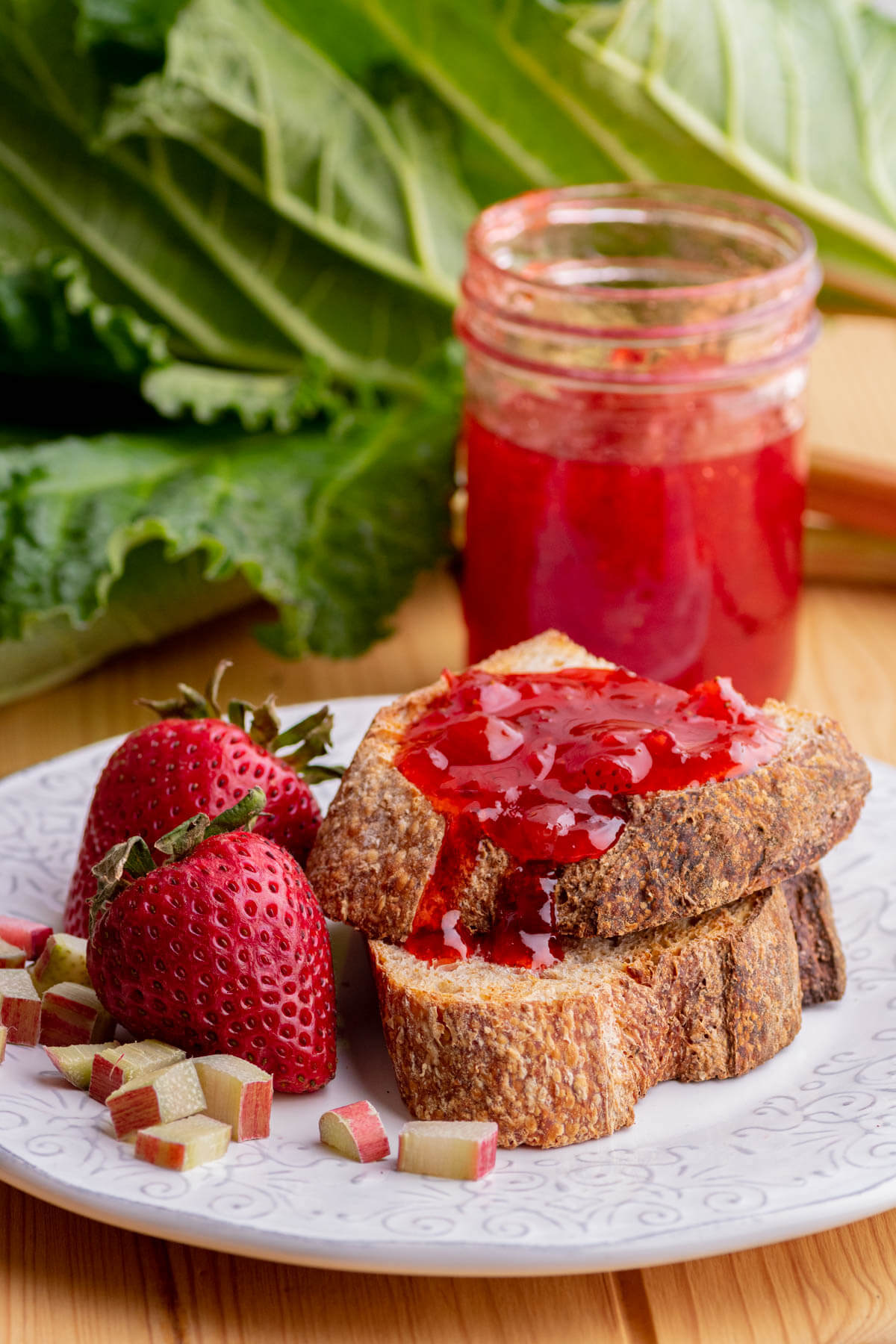 Two sliced of toast slathered in Strawberry Rhubarb Jam with a jar of jam and fresh rhubarb in the background.