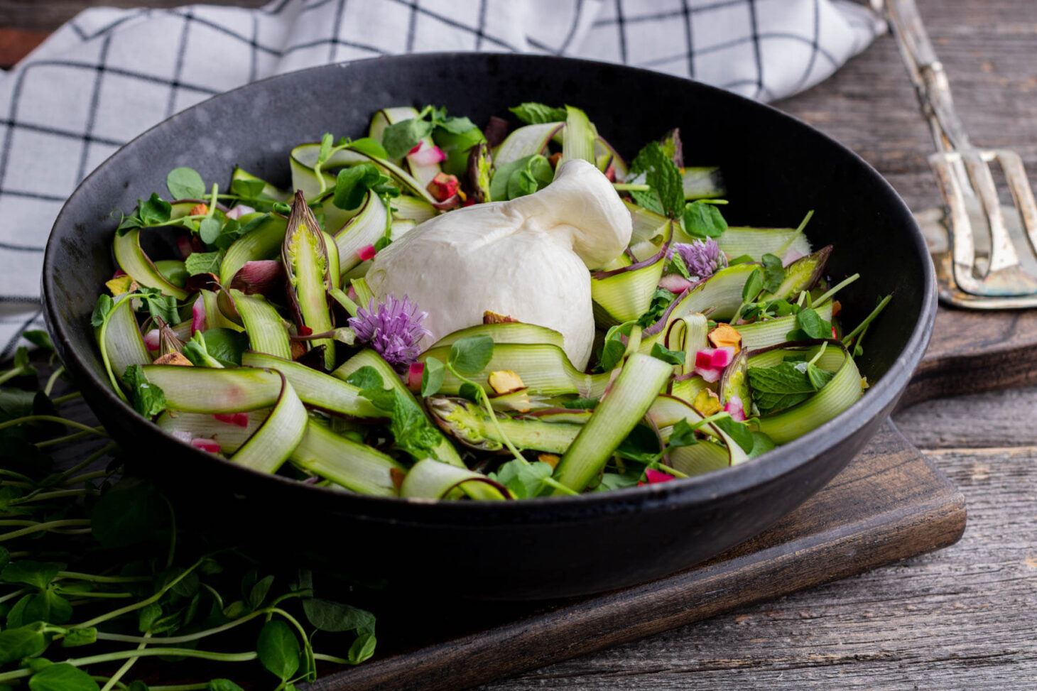 A wide black bowl filled with purple and green shaved asparagus salad, topped with a ball of fresh white burrata.