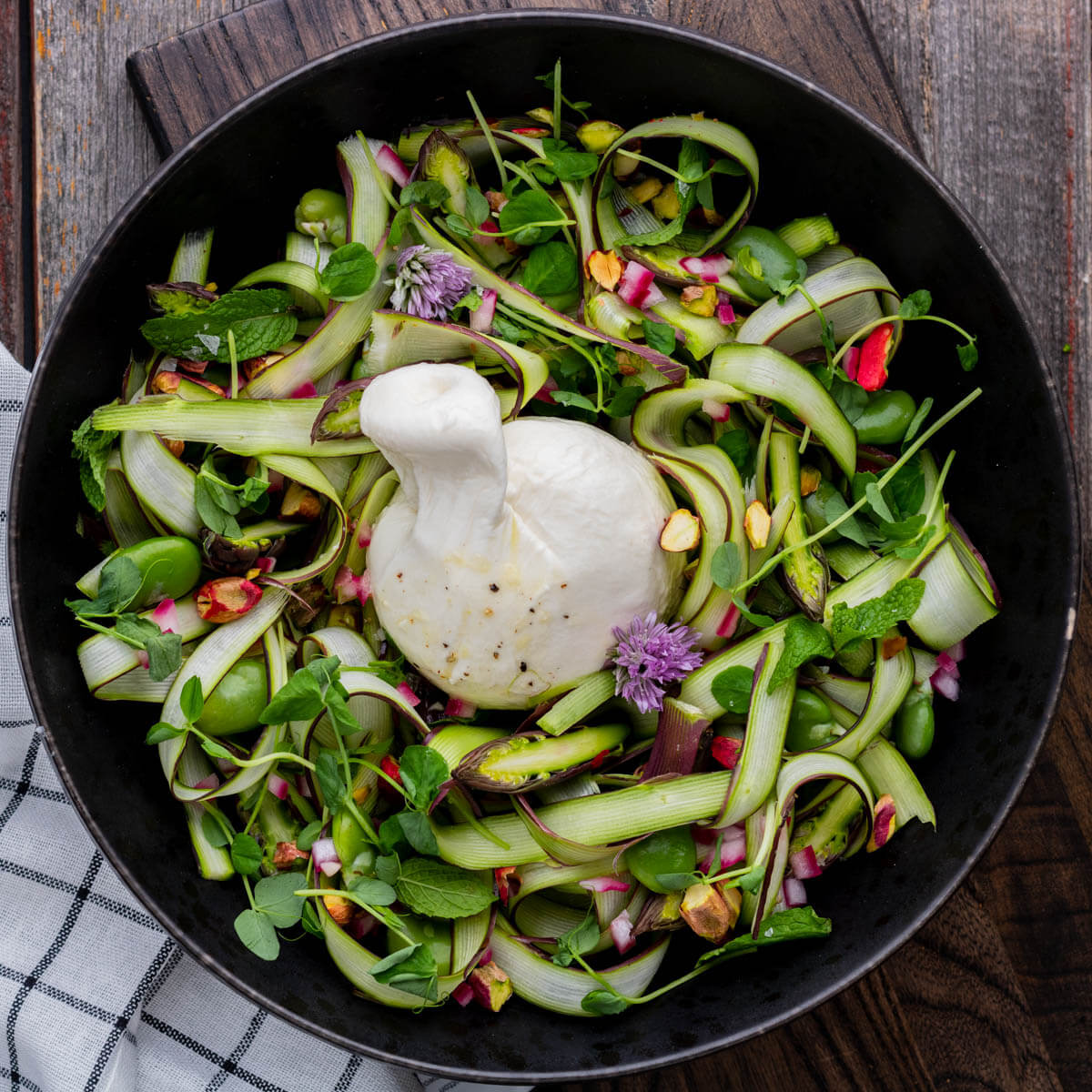 A wide black bowl filled with purple and green shaved asparagus salad, topped with a ball of fresh white burrata.