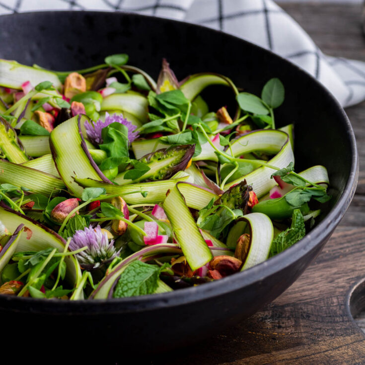 Pinterest image of a wide black bowl filled with purple and green shaved asparagus, pistachios, fava beans, fresh herbs and purple chive flowers. Topped with a white ball of burrata.