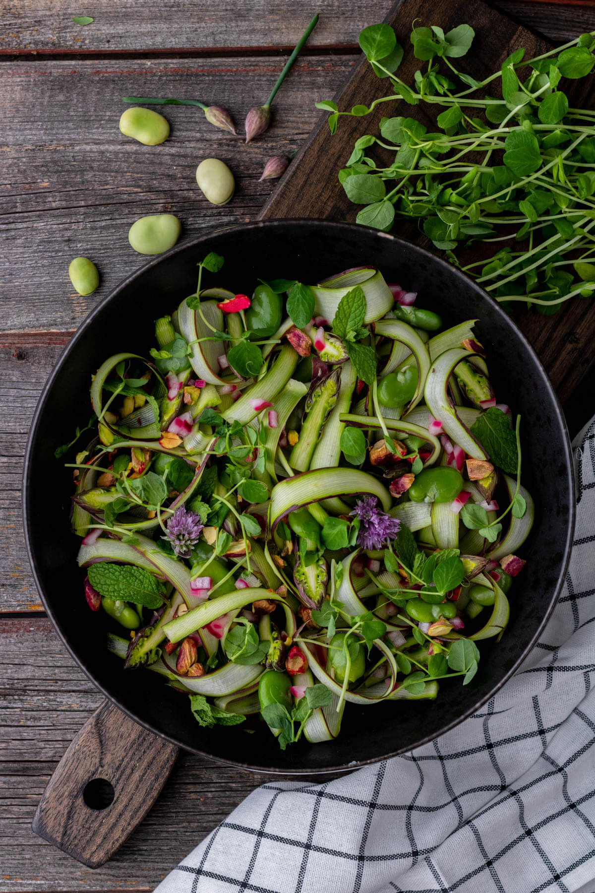 A wide black bowl filled with purple and green shaved asparagus, pistachios, fava beans, fresh herbs and purple chive flowers.