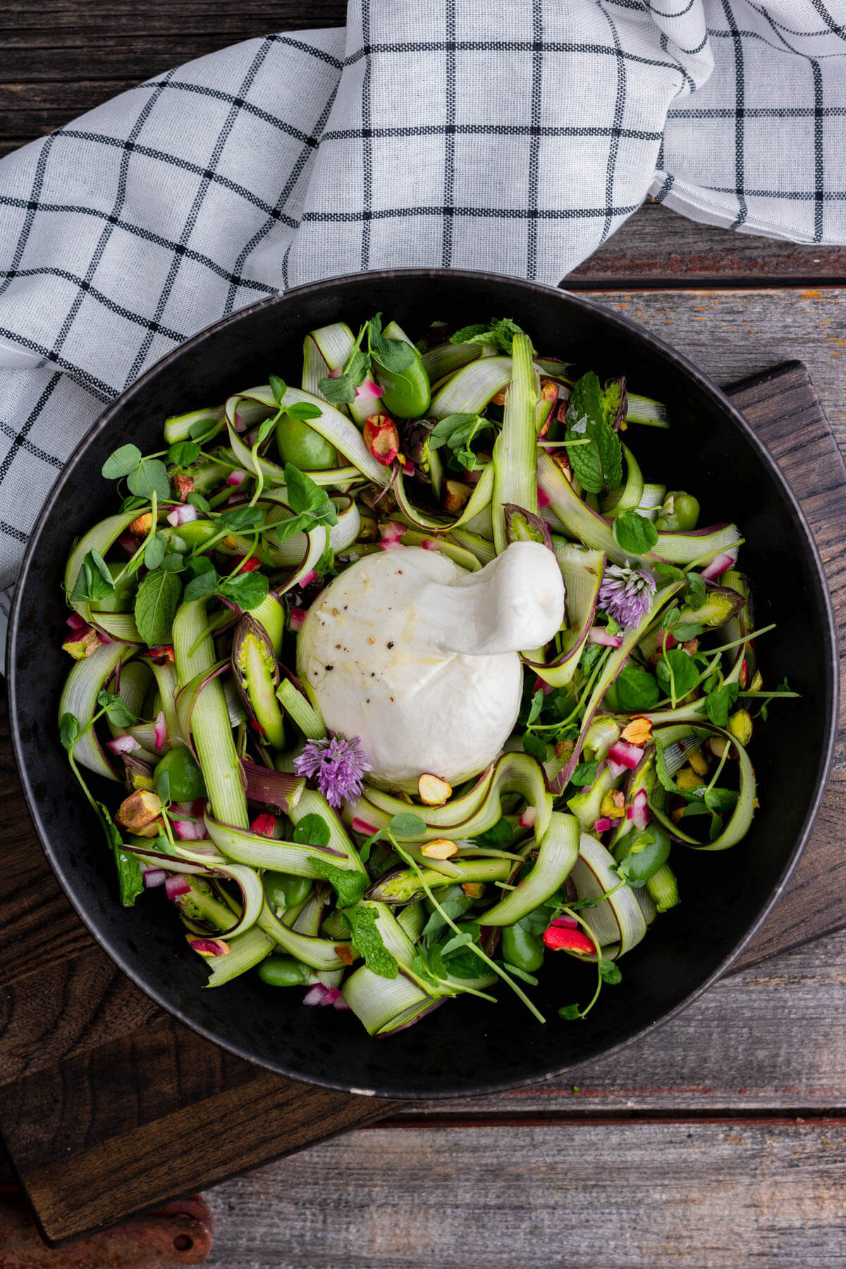 A wide black bowl filled with purple and green shaved asparagus salad, topped with a ball of fresh white burrata. 