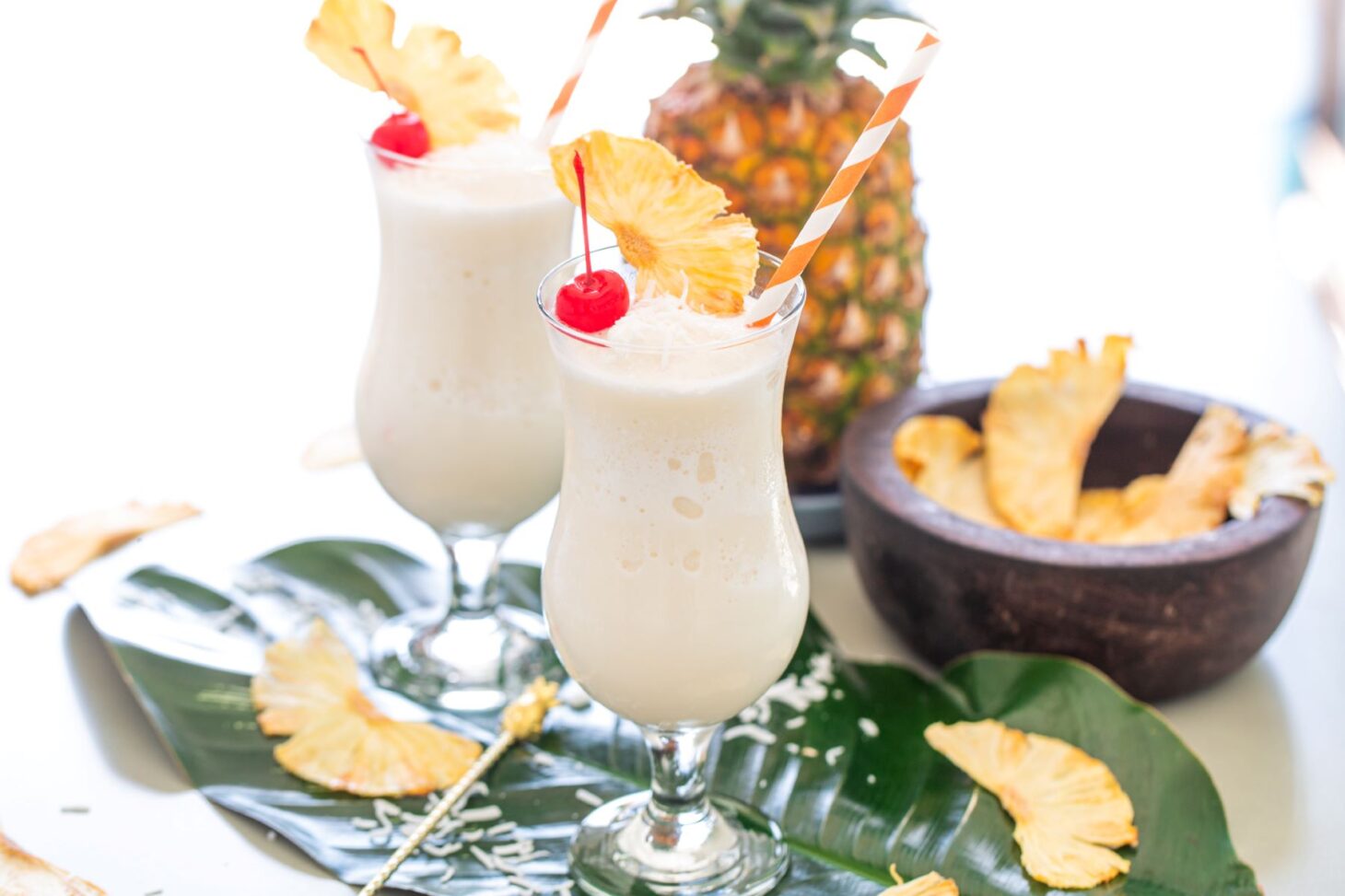 Two pina coladas in hurricane glasses surrounded by pineapple, dried pineapple wedges, and shredded coconut.