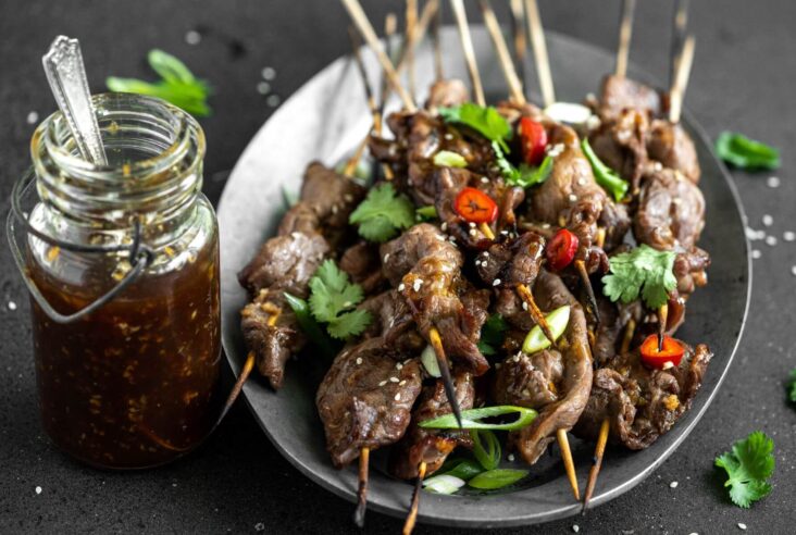 A platter of grilled Teriyaki Beef Sticks garnished with chopped green herbs and red hot peppers. A jar of teriyaki sauce on the side.