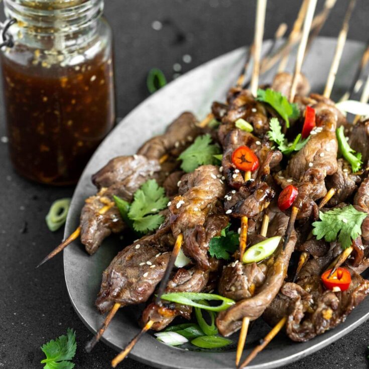 A platter of grilled Teriyaki Beef Sticks garnished with chopped green herbs and red hot peppers. A jar of teriyaki sauce on the side.
