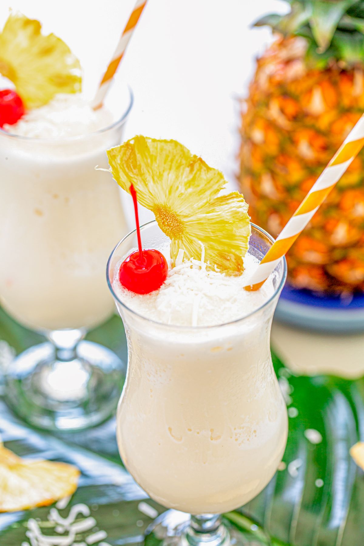 Two pina coladas garnished with a red cherry, dried pineapple wedge, and shredded coconut.