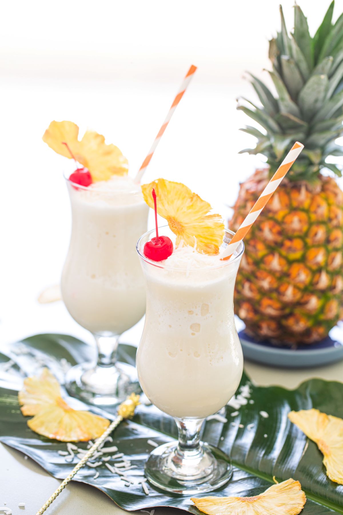 Two pina coladas garnished with a red cherry, dried pineapple wedge, and shredded coconut.