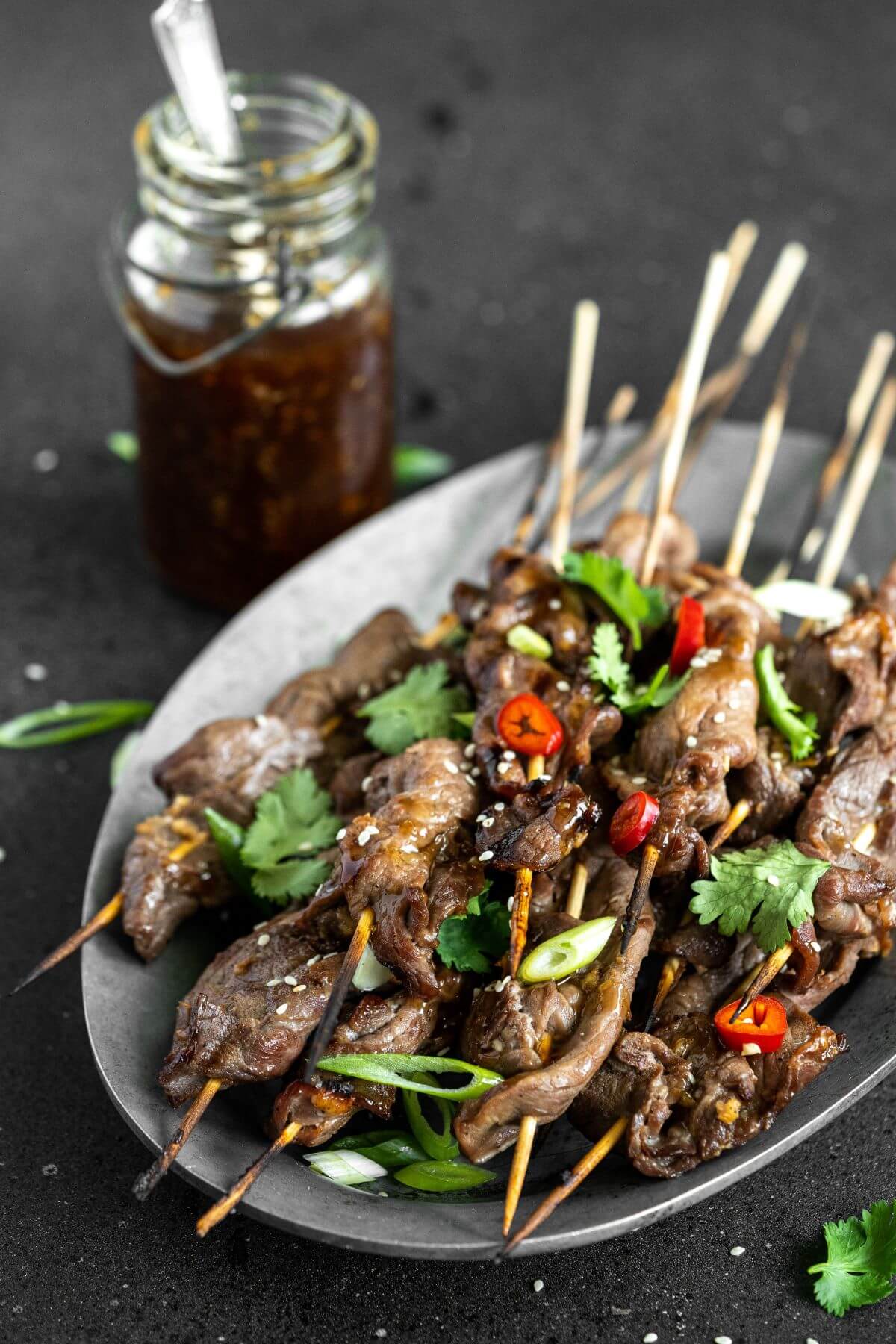 A platter of grilled Teriyaki Beef Sticks garnished with chopped green herbs and red hot peppers. A jar of teriyaki sauce on the side.