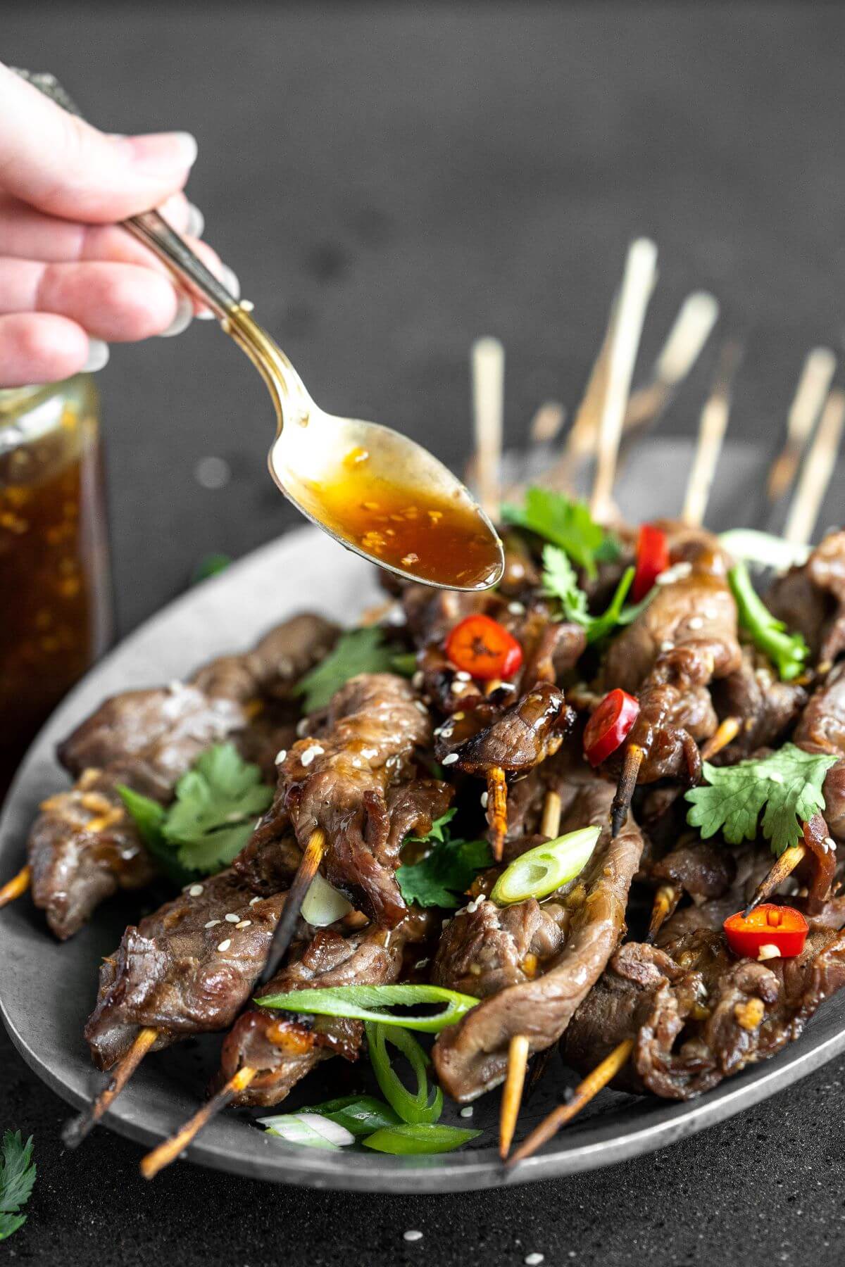 Teriyaki sauce being spooned over a platter of grilled Teriyaki Beef Sticks garnished with chopped green herbs and red hot peppers.
