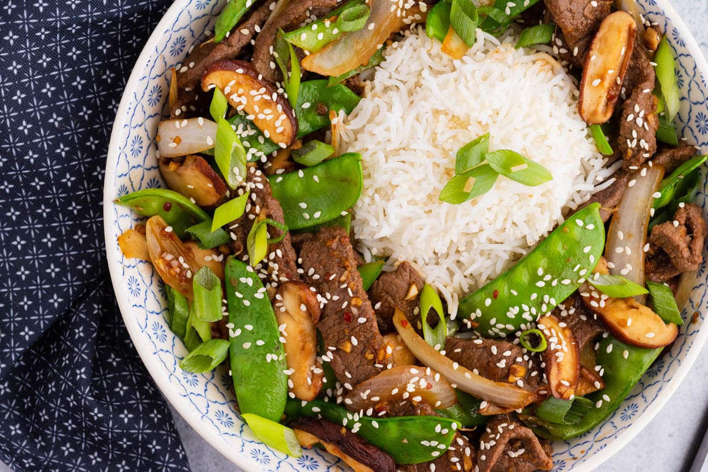 A bowl filled with beef stir fry and snow peas, white rice, shiitake mushrooms topped with sesame seeds and sliced green onions.