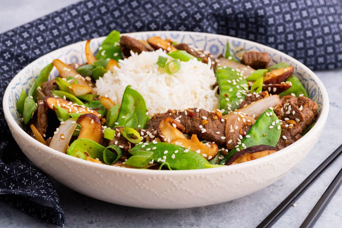 A bowl filled with beef stir fry and snow peas, white rice, shiitake mushrooms topped with sesame seeds and sliced green onions.
