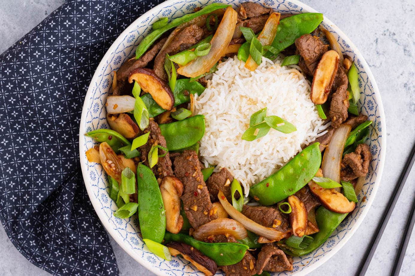 A bowl filled with beef stir fry and snow peas, white rice, shiitake mushrooms.