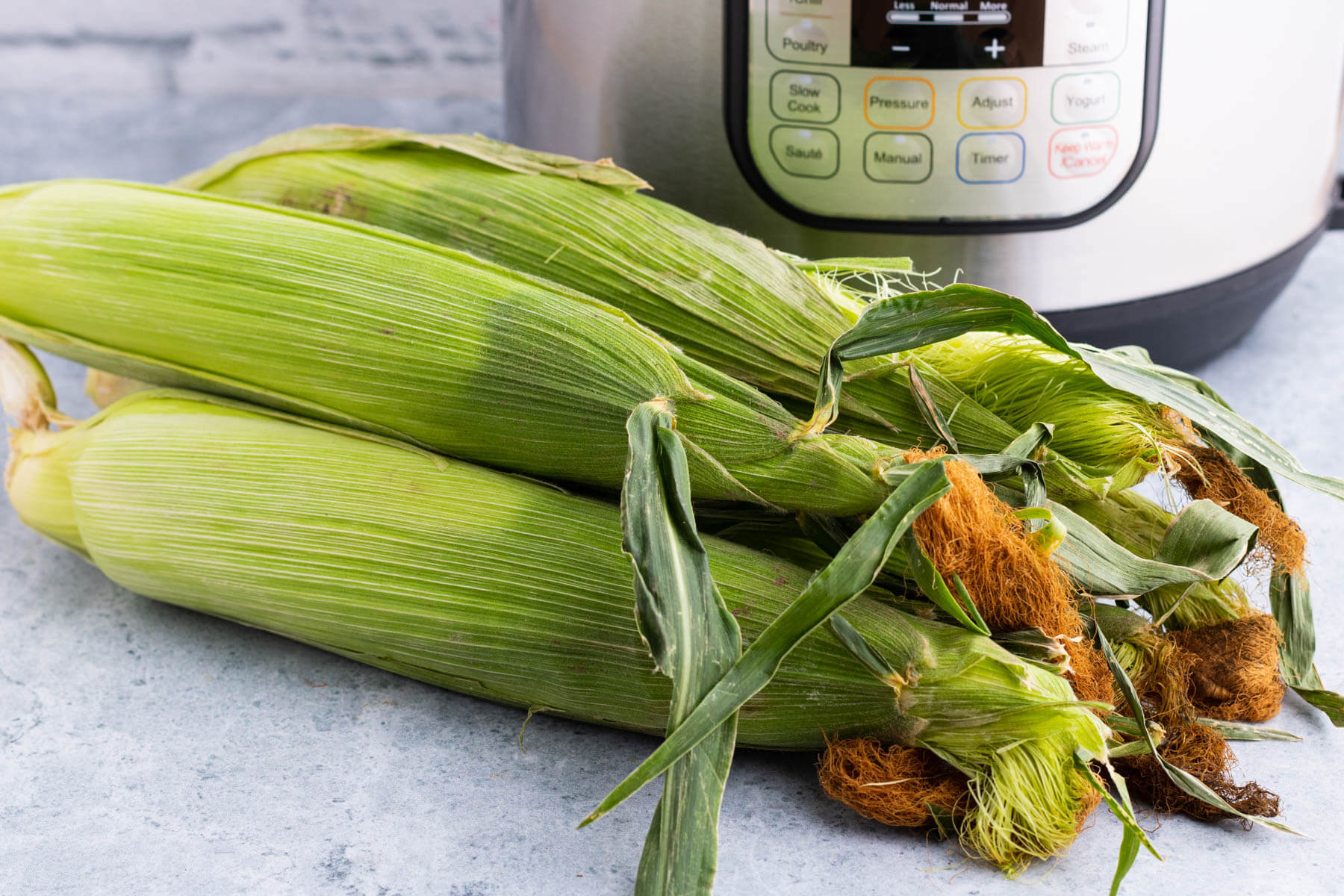 A stack of green ears of corn in front of an Instant Pot.