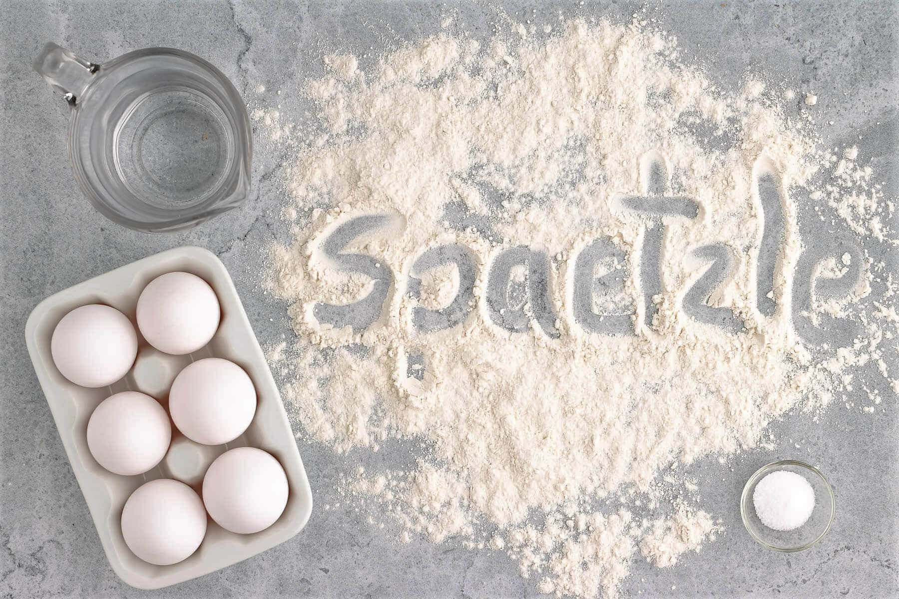 Spilled flour with the word 'spaetzle' written into it. Eggs, salt, and water nearby.