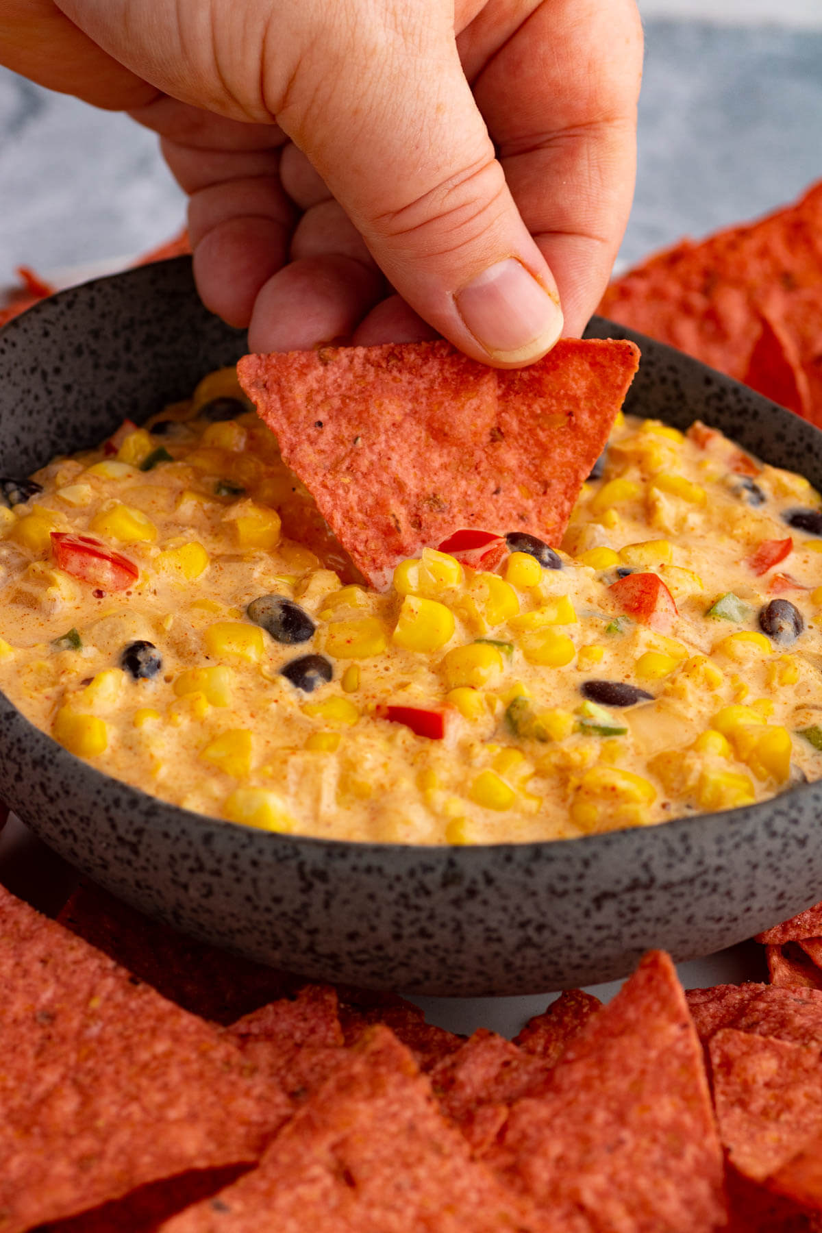 A hand dips a red tortilla chip being dipped in a bowl of creamy corn dip.
