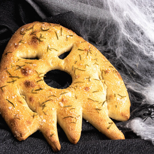 A ghost shaped baked fougasse flat bread on a dark background surrounded by spiderwebs..