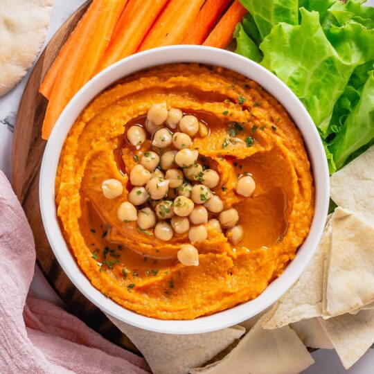 A white bowl of pumpkin hummus topped with chickpeas and olive oil surrounded by pita bread and carrot sticks.