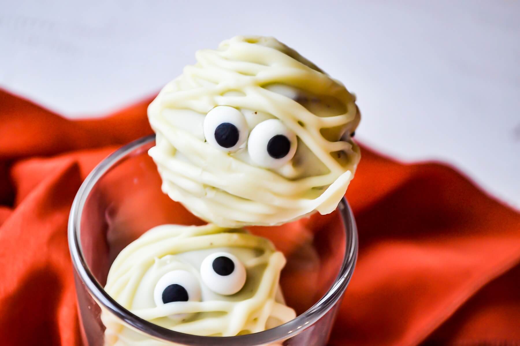 Two white chocolate mummified white chocolate oreo truffles with candy eyes sit in a glass.