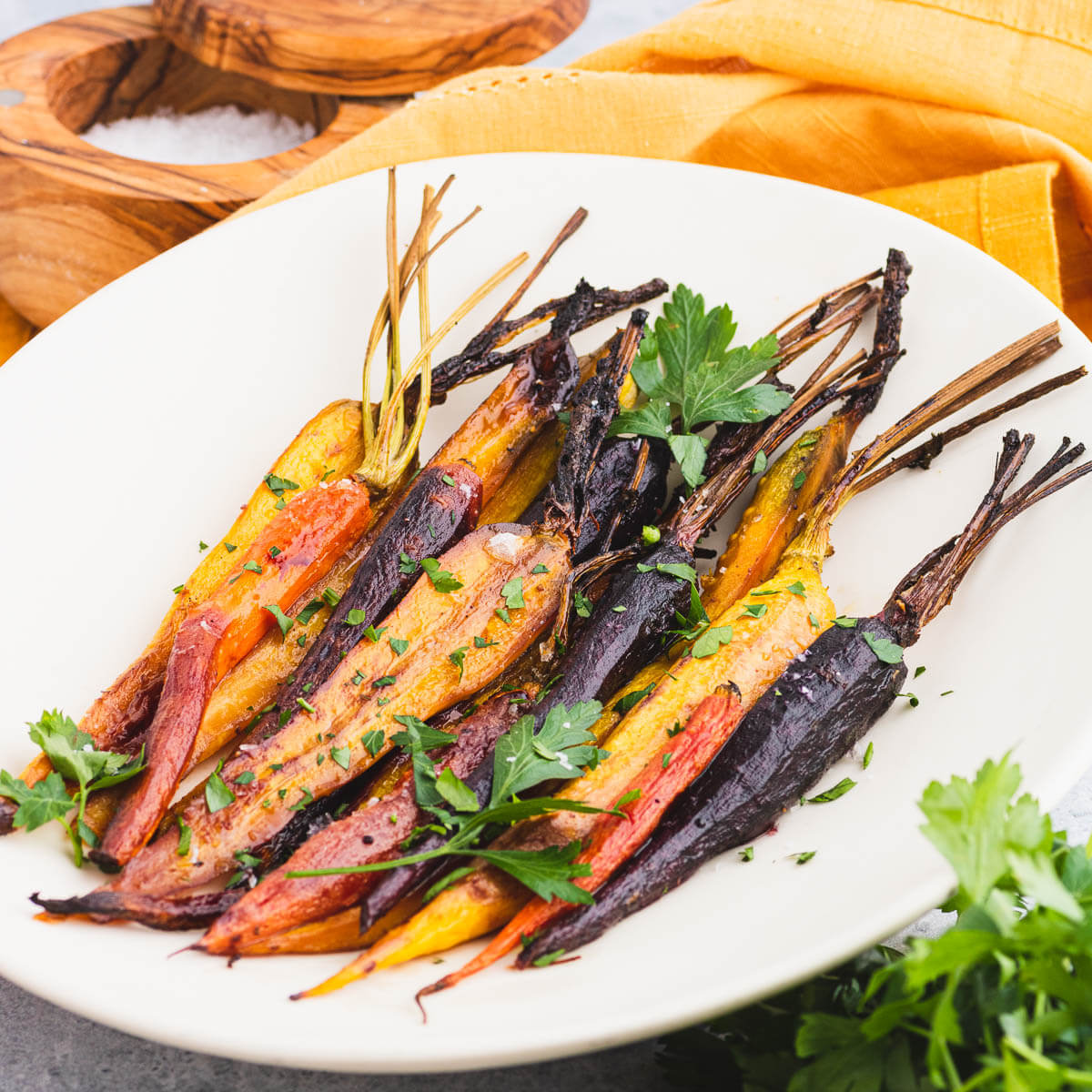 A white oval platter containing colourful maple balsamic roasted carrots garnished with coarse salt and chopped parsley.
