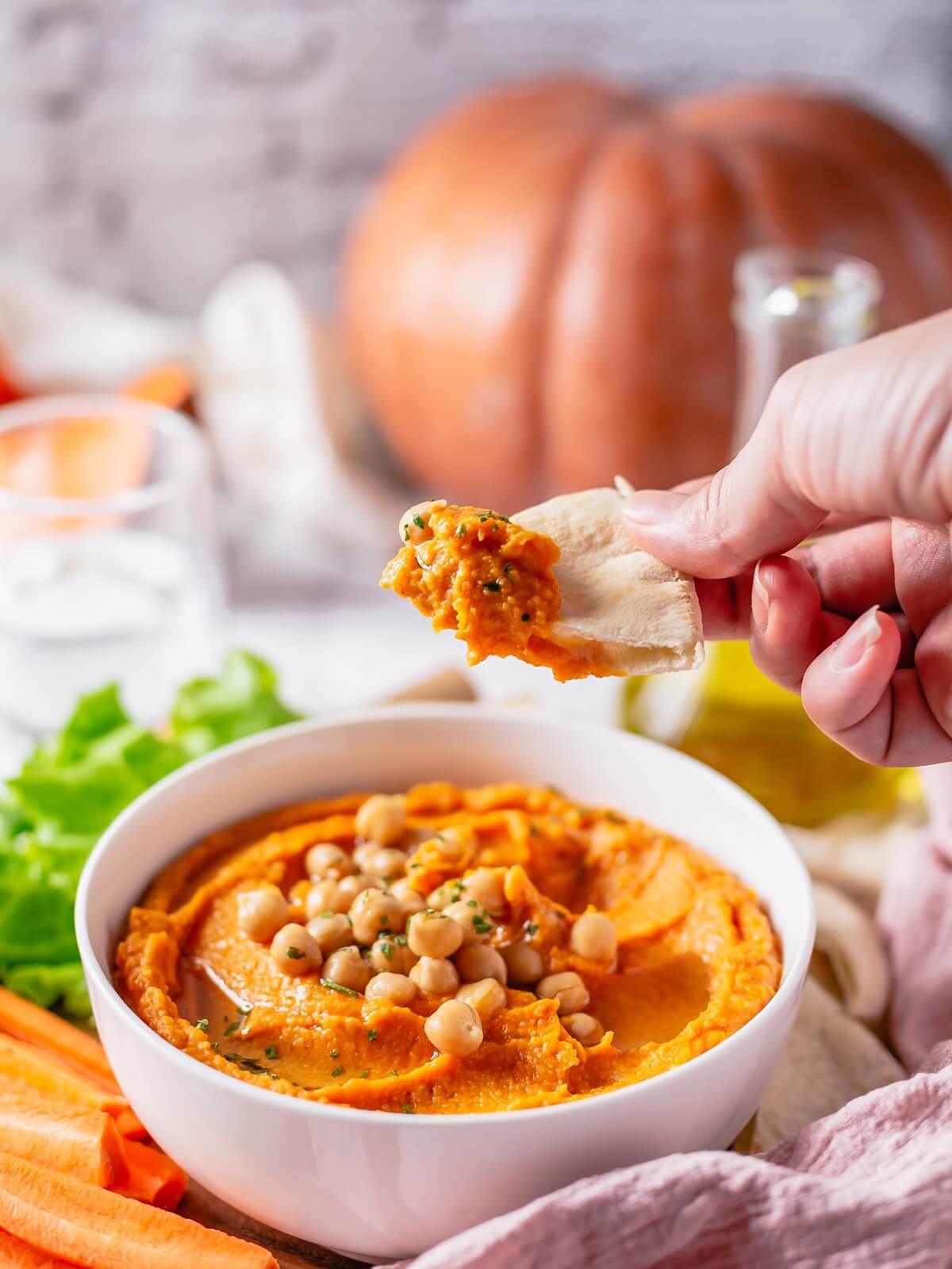A hand holds a triangle of pita bread topped with pumpkin hummus over a bowl of the same.