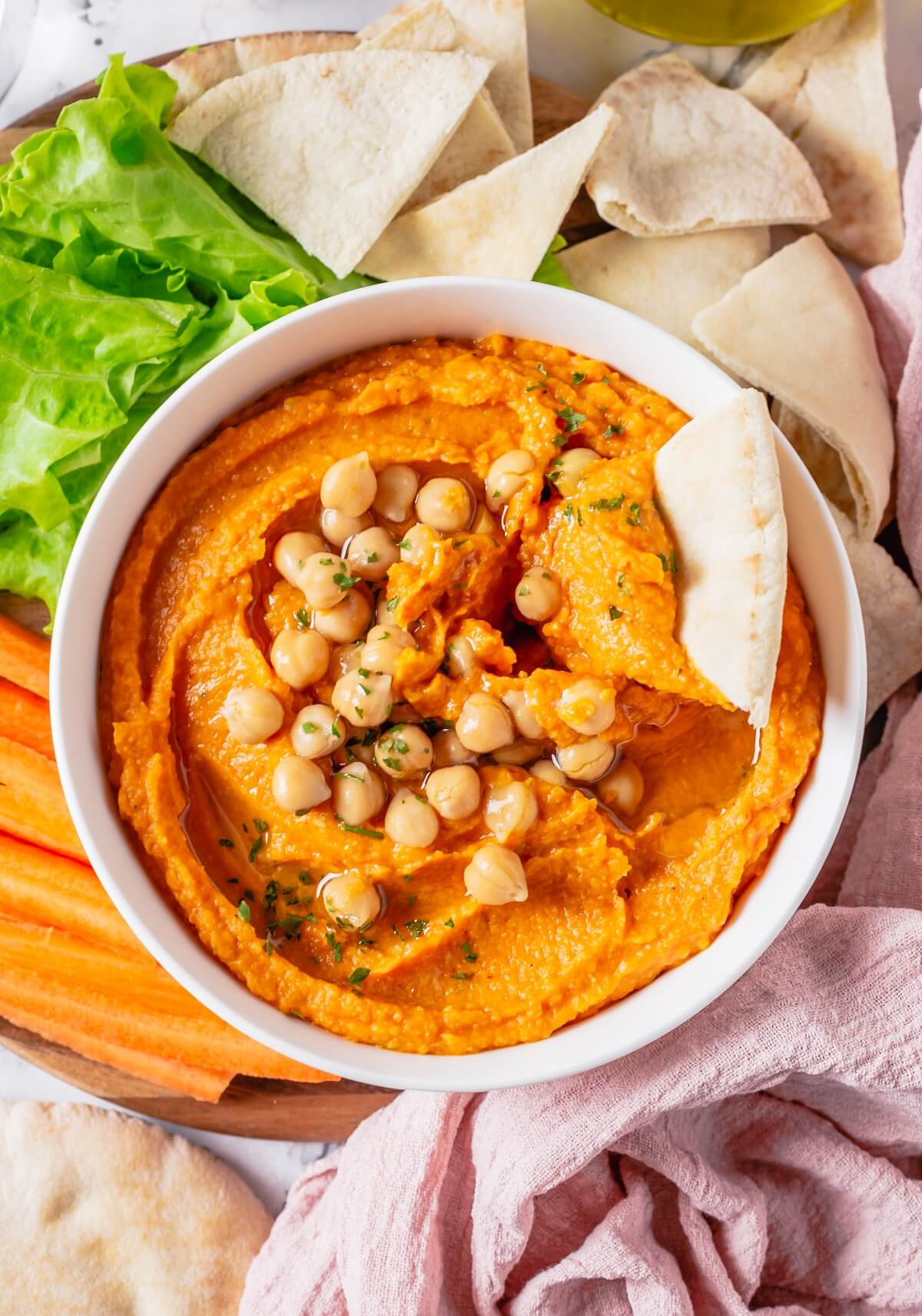 A white bowl of pumpkin hummus topped with chickpeas and olive oil surrounded by pita bread and carrot sticks.