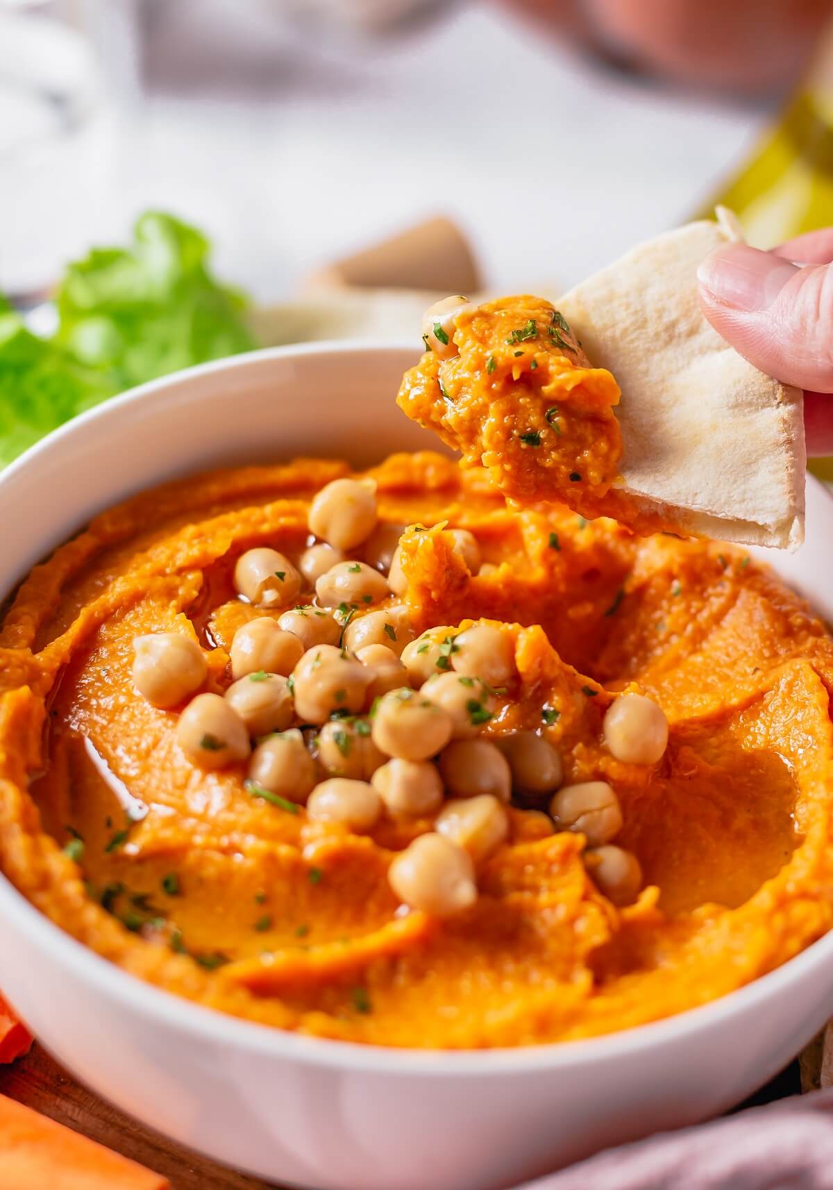 A hand holds a triangle of pita bread topped with pumpkin hummus over a bowl of the same.