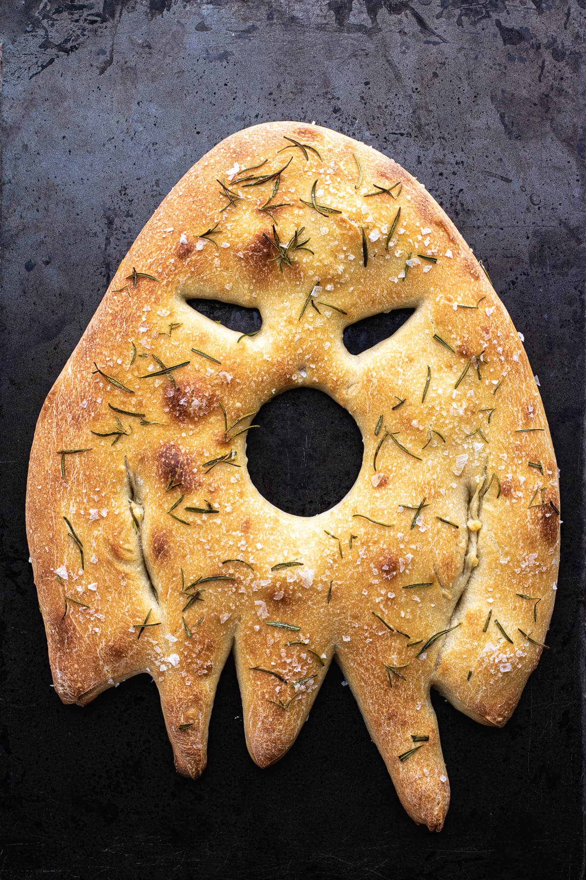 A ghost shaped fougasse flat bread on a dark baking pan.