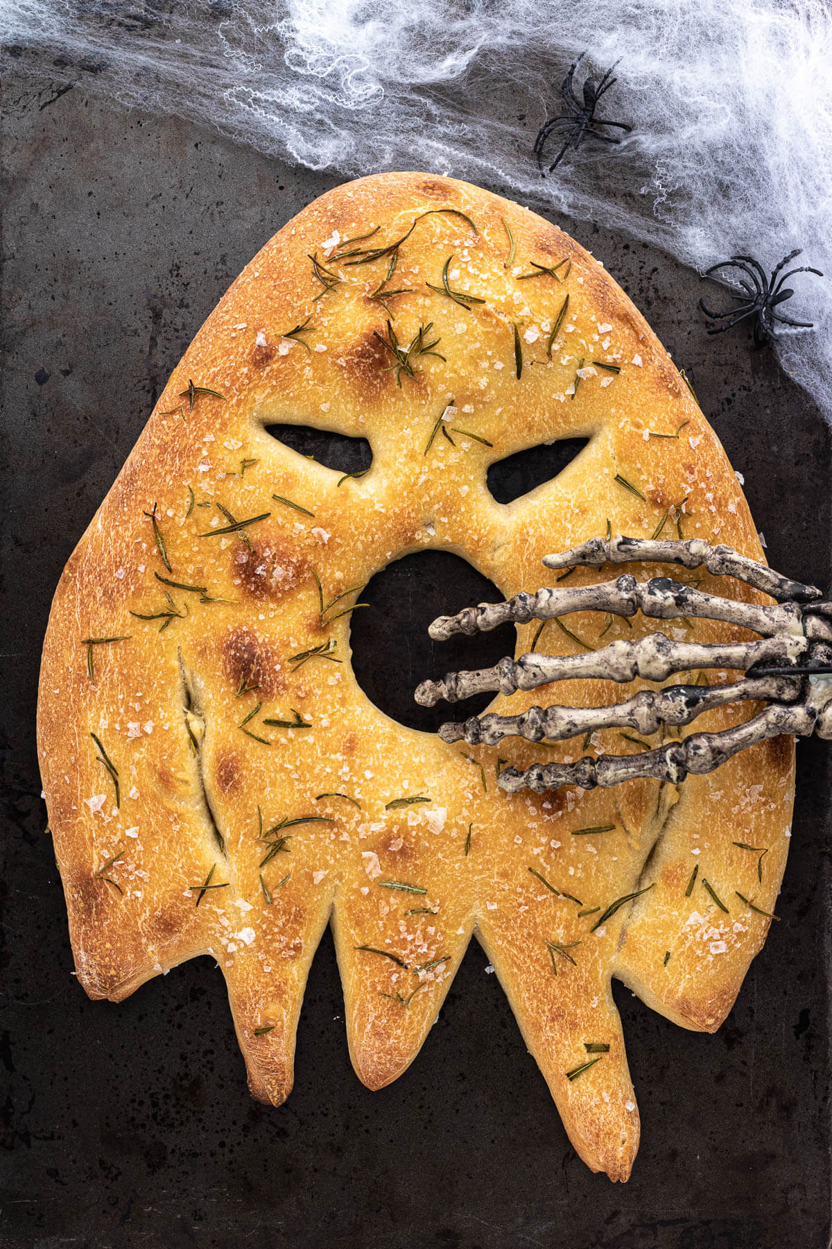 A ghost shaped fougasse flat bread on a dark baking pan with skeleton hand covering the open mouth.