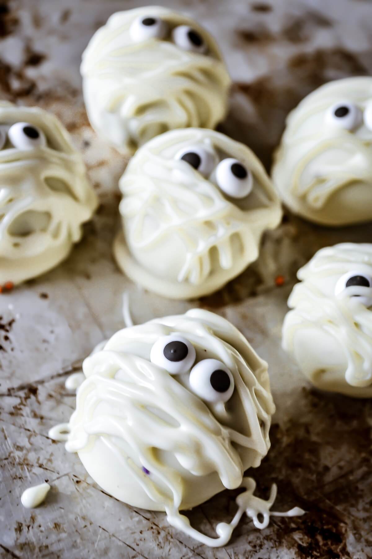 Six Mummified white Oreo Truffle with candy eyes sit on a spattered dark baking sheet.