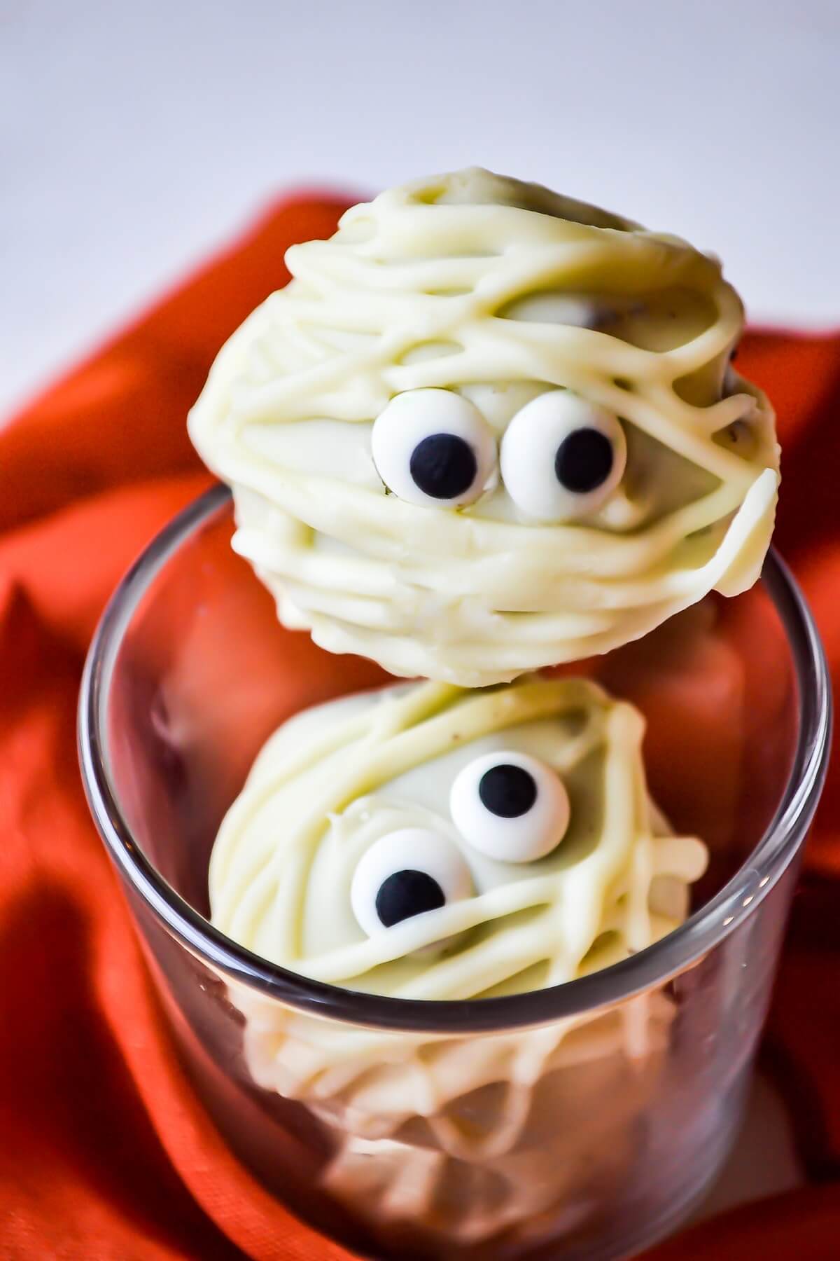 Two white chocolate mummified white chocolate oreo truffles with candy eyes sit in a glass.